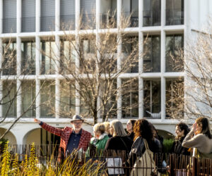 A group of people participates in a walking tour in a lush urban setting with a modernist architectural building in the background. The tour guide, wearing a red checkered jacket and a hat, gestures animatedly while explaining details to the attendees. Bare trees frame the scene, and the atmosphere is lively and engaging, reflecting the exploration of Robin Boyd's architectural neighborhood. Photo: Mike Lam.