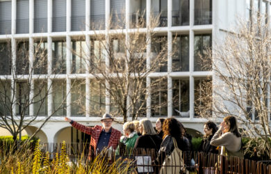 A group of people participates in a walking tour in a lush urban setting with a modernist architectural building in the background. The tour guide, wearing a red checkered jacket and a hat, gestures animatedly while explaining details to the attendees. Bare trees frame the scene, and the atmosphere is lively and engaging, reflecting the exploration of Robin Boyd's architectural neighborhood. Photo: Mike Lam.