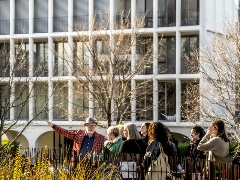 A group of people participates in a walking tour in a lush urban setting with a modernist architectural building in the background. The tour guide, wearing a red checkered jacket and a hat, gestures animatedly while explaining details to the attendees. Bare trees frame the scene, and the atmosphere is lively and engaging, reflecting the exploration of Robin Boyd's architectural neighborhood. Photo: Mike Lam.