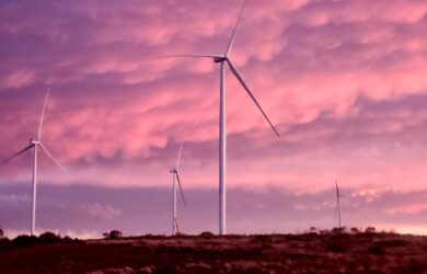 A wind farm set against a stunning pink and purple sky, with multiple wind turbines standing tall on the horizon. The colors of the sunset create a surreal and atmospheric scene.