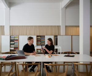 Two designers sitting at a modern workspace, discussing materials and samples at a table with bookshelves in the background.