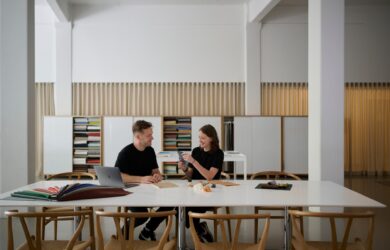 Two designers sitting at a modern workspace, discussing materials and samples at a table with bookshelves in the background.