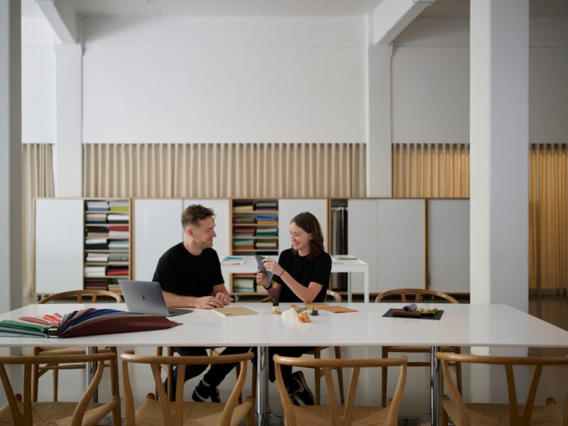 Two designers sitting at a modern workspace, discussing materials and samples at a table with bookshelves in the background.