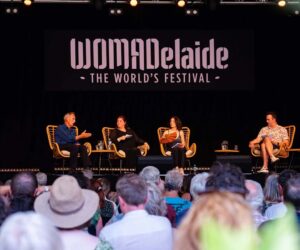 A panel discussion at WOMADelaide, "The World's Festival," featuring four speakers seated on a stage with a black backdrop displaying the festival's logo.