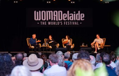 A panel discussion at WOMADelaide, "The World's Festival," featuring four speakers seated on a stage with a black backdrop displaying the festival's logo.