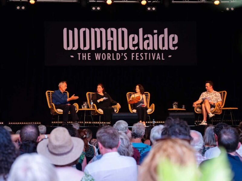 A panel discussion at WOMADelaide, "The World's Festival," featuring four speakers seated on a stage with a black backdrop displaying the festival's logo.