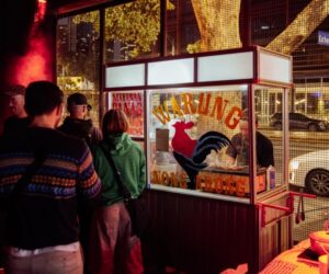 People gathered at a street food stall at night, with food preparation visible inside a lit kiosk.