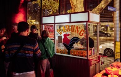 People gathered at a street food stall at night, with food preparation visible inside a lit kiosk.