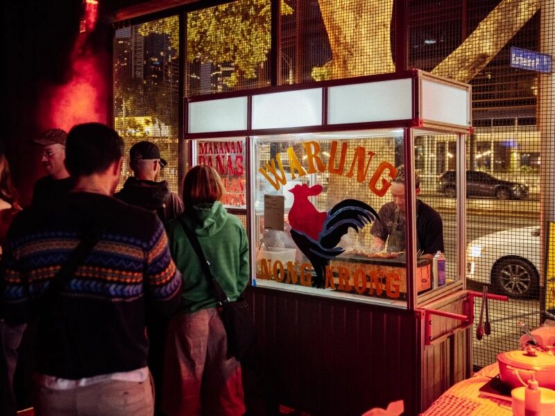 People gathered at a street food stall at night, with food preparation visible inside a lit kiosk.