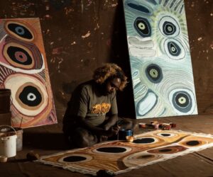Corban Clause Williams painting a large canvas on the floor inside a studio, with two completed paintings propped against a wall behind him.
