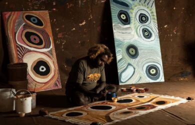 Corban Clause Williams painting a large canvas on the floor inside a studio, with two completed paintings propped against a wall behind him.