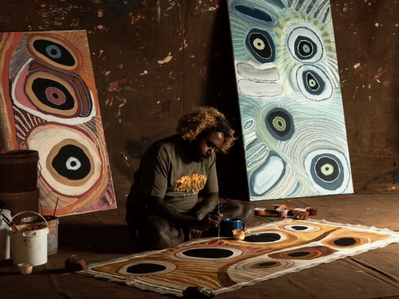 Corban Clause Williams painting a large canvas on the floor inside a studio, with two completed paintings propped against a wall behind him.