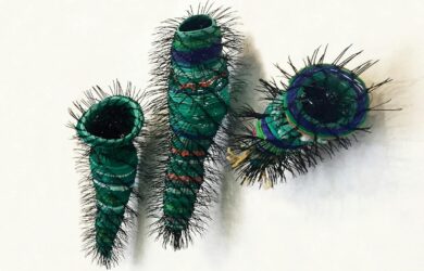 Three sculptural vessels made from layered recycled plastic in green and blue tones, wrapped with fine black spines, mounted on a white wall.